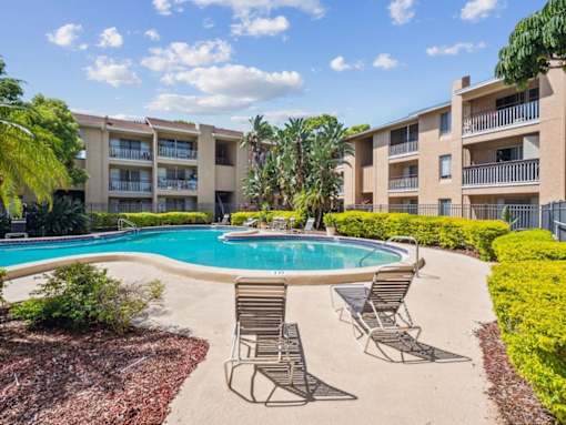 A swimming pool surrounded by chairs and plants in front of apartment buildings.