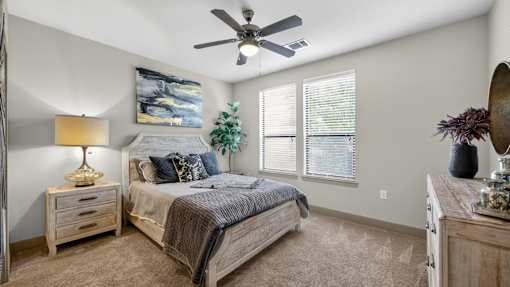 A bedroom with a bed, a dresser, a nightstand, a ceiling fan, and a painting on the wall.