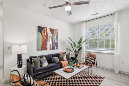 A living room with a black couch, a painting on the wall, a ceiling fan, and a large window with a plant in front of it.