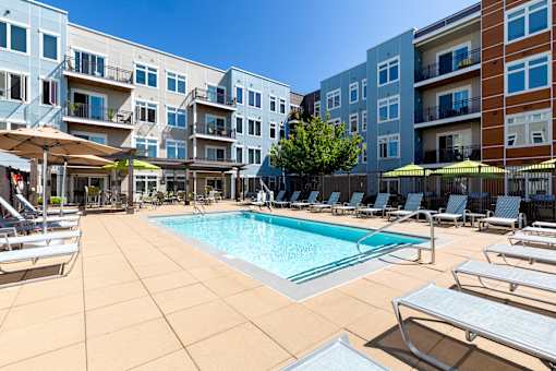 A pool surrounded by sun loungers and umbrellas in front of apartment buildings.