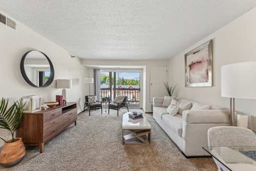 A living room with a white couch, a wooden dresser, a coffee table, and a large window with a view of the outside.