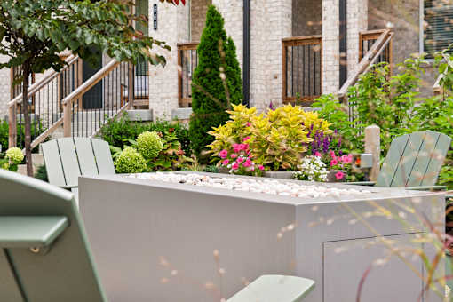 A white bench is in front of a white wall with a tree and flowers in the background.
