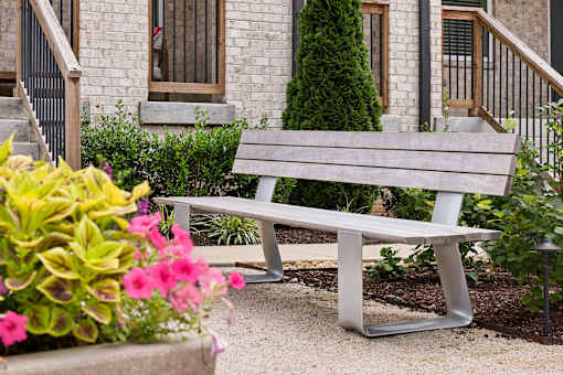 A grey bench is in front of a building with a tree and flowers.