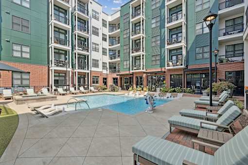 A large swimming pool surrounded by lounge chairs and umbrellas in front of apartment buildings.