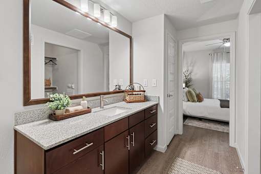 A bathroom with a vanity, mirror, and a bedroom in the background.