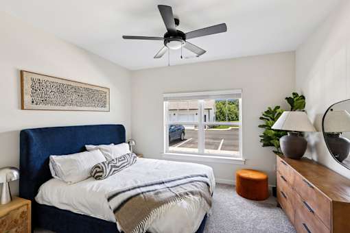 A bedroom with a bed, a ceiling fan, and a window overlooking a parking lot.