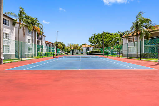 A tennis court with a red surface and a green fence surrounding it.