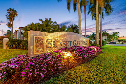 Sunset Gardens sign with flowers in front and palm trees in the background.