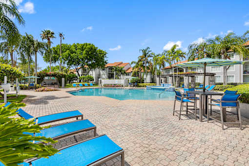 A pool surrounded by palm trees and a patio area with chairs and a table.