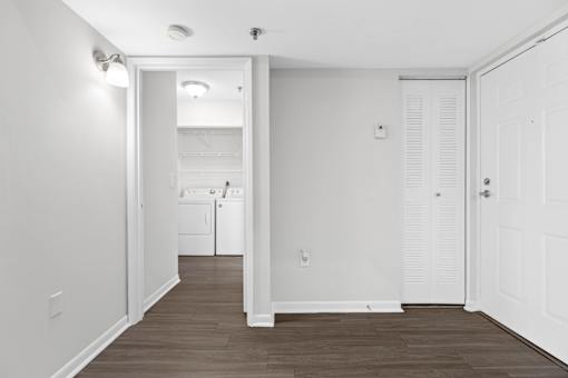 A white hallway with a door on the right and a kitchen visible through the doorway.