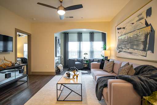 Apartments for Rent in West Nashville, TN - The Summit at Nashville West - Living Room with Sofa, Coffee Table, Ceiling Fan, Wall-Mounted Television, and Bright Window Nook with Chairs.