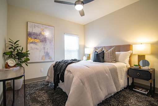 West Nashville, TN, Apartments for Rent - The Summit at Nashville West - Bedroom with Upholstered Headboard, Plush Bedding, Ceiling Fan, Nightstands, and Soft Lighting Beside Window.