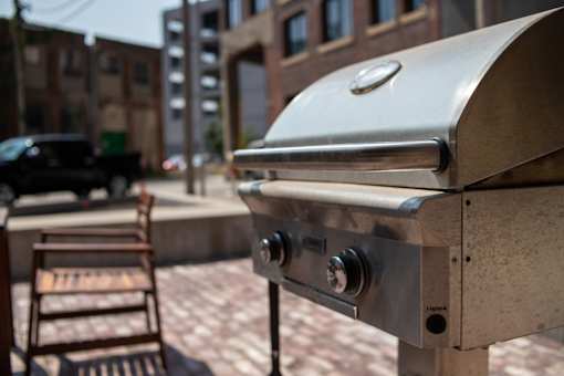 A silver grill with a lid is in the foreground with a chair and a building in the background.