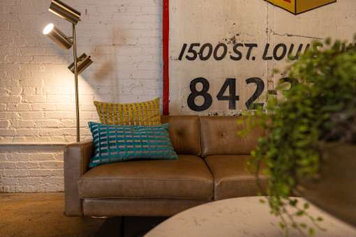 A brown couch with a blue pillow and a yellow pillow is in front of a white brick wall with a sign that says 1500 St. Louis 842.