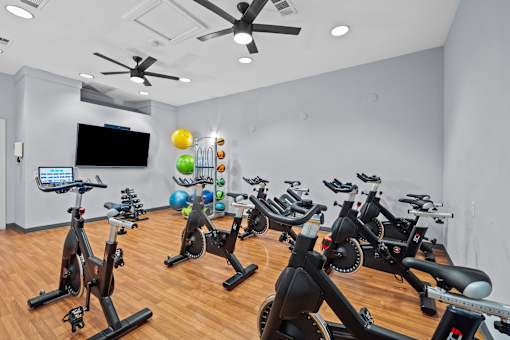 A room with a white wall and a ceiling fan with a row of stationary bikes in front of it.