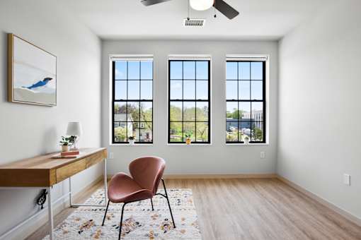 A room with a chair and a table in front of three windows.