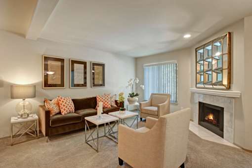 A cozy living room here at Windsong featuring a warm brown sofa with orange accent pillows, beige armchairs, and a sleek mirrored fireplace. Bright, inviting space with soft carpet, elegant side tables, and welcoming natural light.