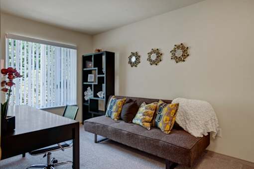 A bright office space here at Windsong featuring a cozy gray sofa with colorful accent pillows, sleek desk, and modern shelving. Large window with vertical blinds brings in natural light, creating a comfortable and inviting workspace.