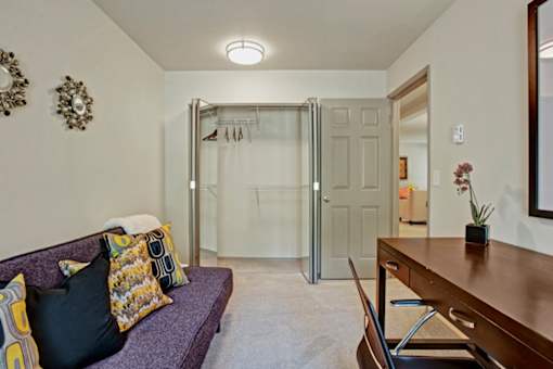 A versatile office space here at Windsong featuring a cozy sofa with colorful accent pillows, spacious desk, and open closet. Neutral tones and soft lighting create a comfortable, welcoming area for work or relaxation.