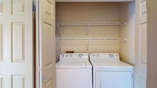 A laundry area here at Windsong featuring full-size washer and dryer units, open shelving for storage, and a clean, organized layout. Bright finishes and convenient design make everyday laundry simple and efficient.