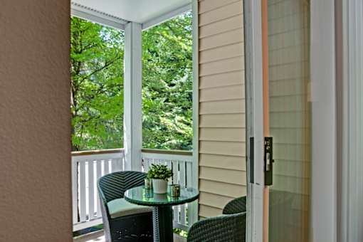 A private balcony here at Windsong featuring a cozy bistro table with two woven chairs, surrounded by lush green tree views. Bright and peaceful, this inviting outdoor space is perfect for relaxing or enjoying fresh air.
