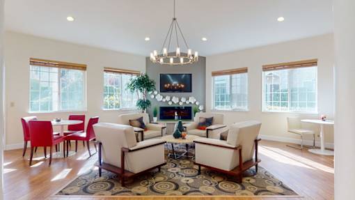 A resident lounge here at Windsong featuring bright windows, cozy armchairs, and a fireplace with mounted TV. Warm and welcoming, this stylish space includes a patterned rug, round chandelier, and inviting seating areas perfect for relaxing or gathering.