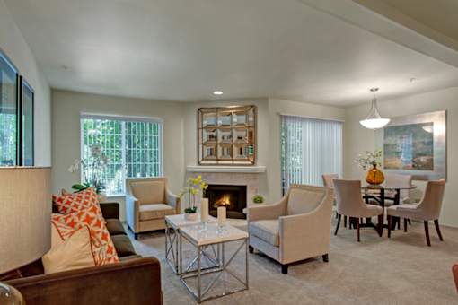 A bright living and dining area here at Windsong featuring a brown sofa with orange accent pillows, beige armchairs, and a mirrored fireplace. Welcoming space with soft carpet, elegant glass table, and large windows bringing natural light.