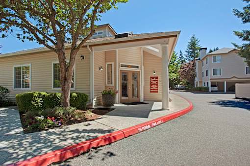 A leasing office exterior here at Windsong featuring a welcoming covered entrance, clean siding, landscaped greenery, and a curved driveway, creating an inviting and well-maintained community arrival experience.