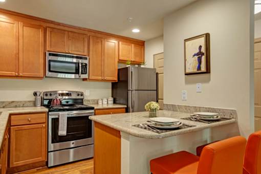 A cozy kitchen here at Windsong featuring warm wood cabinetry, stainless steel appliances, and bright orange barstools. Welcoming layout with a breakfast bar, clean finishes, and inviting, functional design.