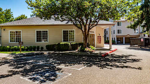 A leasing office exterior here at Windsong featuring a charming single-story building with a covered entry, mature shade tree, and manicured landscaping. This inviting and well-kept space offers convenient access and a welcoming community atmosphere.