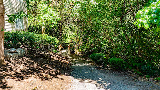 A wooded walking path here at Windsong featuring lush greenery, mature trees, and a charming wooden bridge. This peaceful and scenic setting offers shaded trails and a tranquil atmosphere perfect for relaxing outdoor strolls.