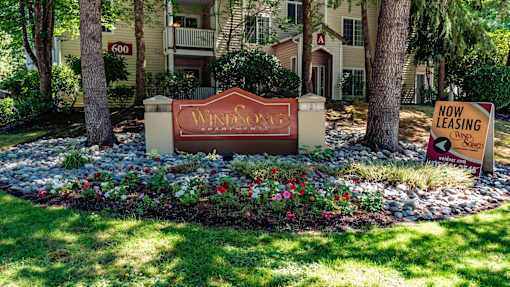 A community entrance sign here at Windsong featuring a warm, welcoming monument sign surrounded by colorful flowers, decorative rocks, mature trees, and lush green landscaping with apartment buildings in the background.