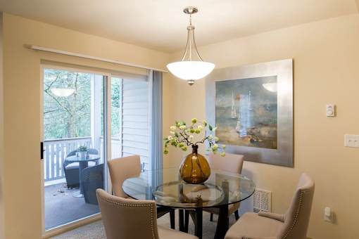 A bright dining area here at Windsong featuring a glass table, beige upholstered chairs, and an amber centerpiece vase. Welcoming layout with sliding door access to a private balcony and soft, natural light.