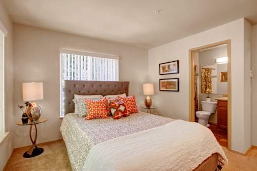 A cozy bedroom here at Windsong featuring a tufted headboard bed, orange accent pillows, and matching bedside lamps. Bright, welcoming layout with soft carpet, neutral tones, and convenient ensuite bathroom access.
