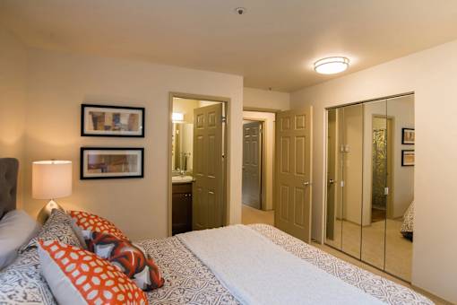 A welcoming bedroom here at Windsong featuring mirrored closet doors, soft carpet, and bright neutral walls. Cozy bed with patterned bedding and orange accent pillows adds warmth, while open doorways provide convenient access to bathroom and storage areas.