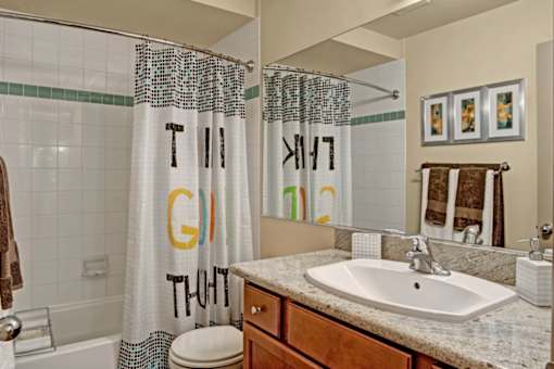 A clean bathroom here at Windsong featuring a granite vanity, large mirror, and bright lighting. Neutral tile shower with patterned curtain and warm wood cabinetry create a comfortable, inviting space.