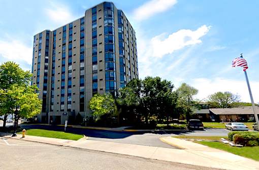 A tall building with a flag on top is surrounded by trees and a street in front.