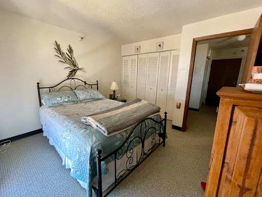 A bed with a metal frame and a blue patterned bedspread sits in a room with a wooden cabinet and a doorway leading to another room.