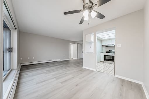A spacious room with a ceiling fan and light, hardwood floors, and a kitchen visible through an open door.