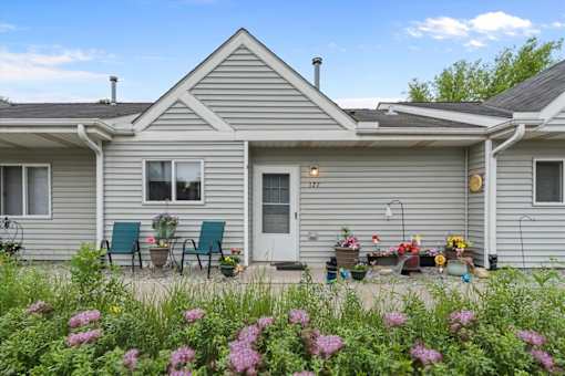 A house with a grey exterior and a white door is surrounded by a garden with pink flowers.