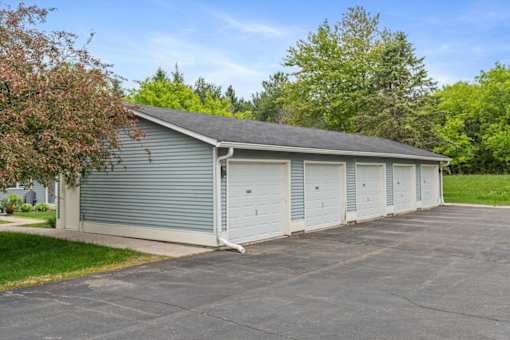 A two-car garage with a grey roof and white doors is surrounded by a paved driveway and greenery.