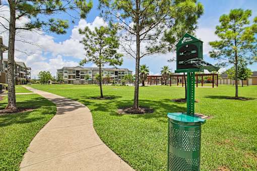A green trash can is in the foreground of a grassy area with a pathway and trees.