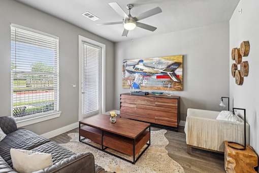 A living room with a grey couch, a coffee table, a painting, a fan, and a window with blinds.