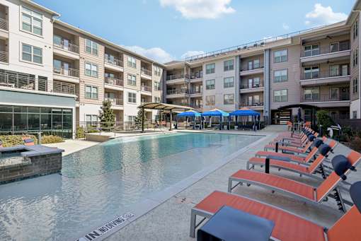 A pool surrounded by sun loungers and umbrellas in front of apartment buildings.
