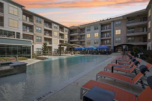 A swimming pool surrounded by sun loungers and umbrellas in front of apartment buildings.