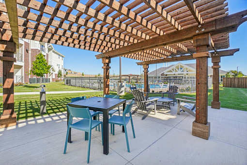 A wooden pergola with a table and chairs is in the foreground of a patio.
