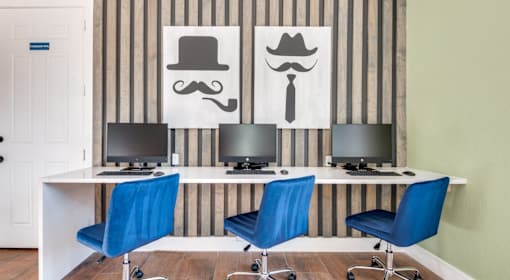 A room with striped wallpaper and a desk with three computers and three blue chairs.