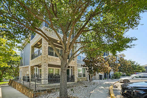 A tree in front of a two-story building with a stone wall and a black car parked on the street.