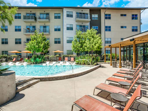 A large swimming pool surrounded by lounge chairs and umbrellas in front of a multi-story apartment building.
