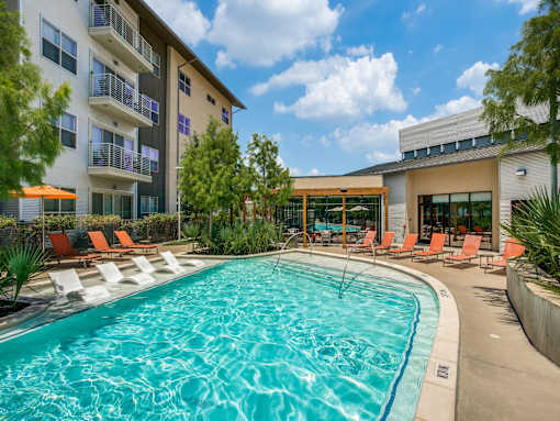 A large swimming pool in front of a building with orange lounge chairs around it.